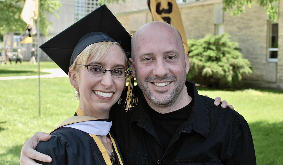 "A woman wearing a graduation cap and gown smiles as she stands next to her brother Patrick smiling and holding her shoulder"